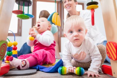 Cute baby girl showing progress and curiosity Cute baby girl showing progress and curiosity by trying to reach multicolored wooden toys, while sitting on the floor with her mother and a family friend